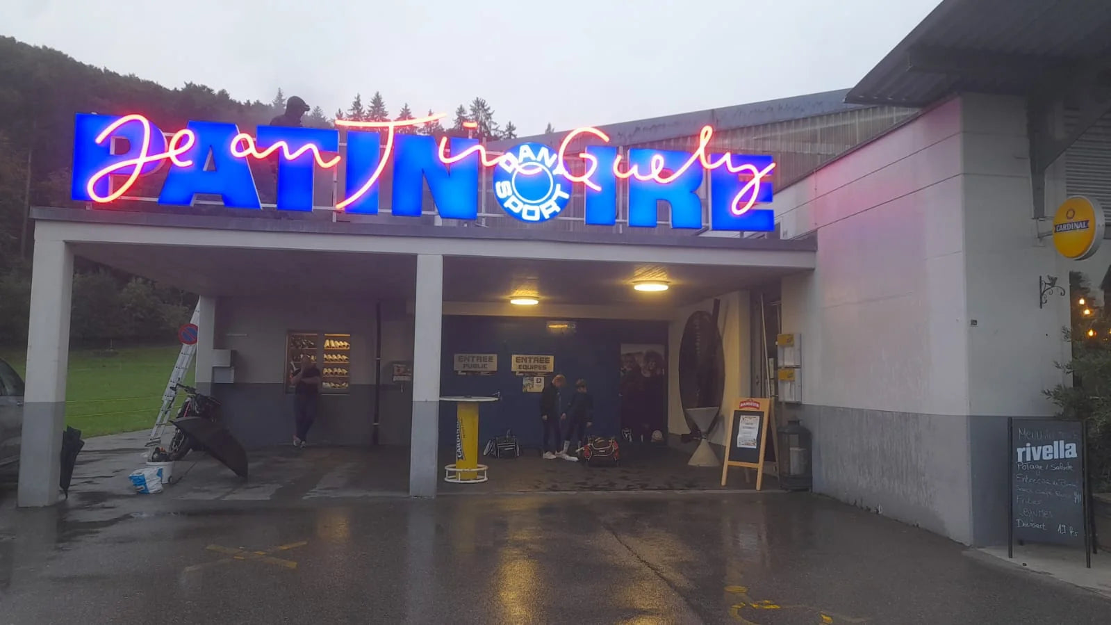 Entrance to the Jean Tinguely ice rink in Marly at dusk, with its new blue sign and red script illuminated.