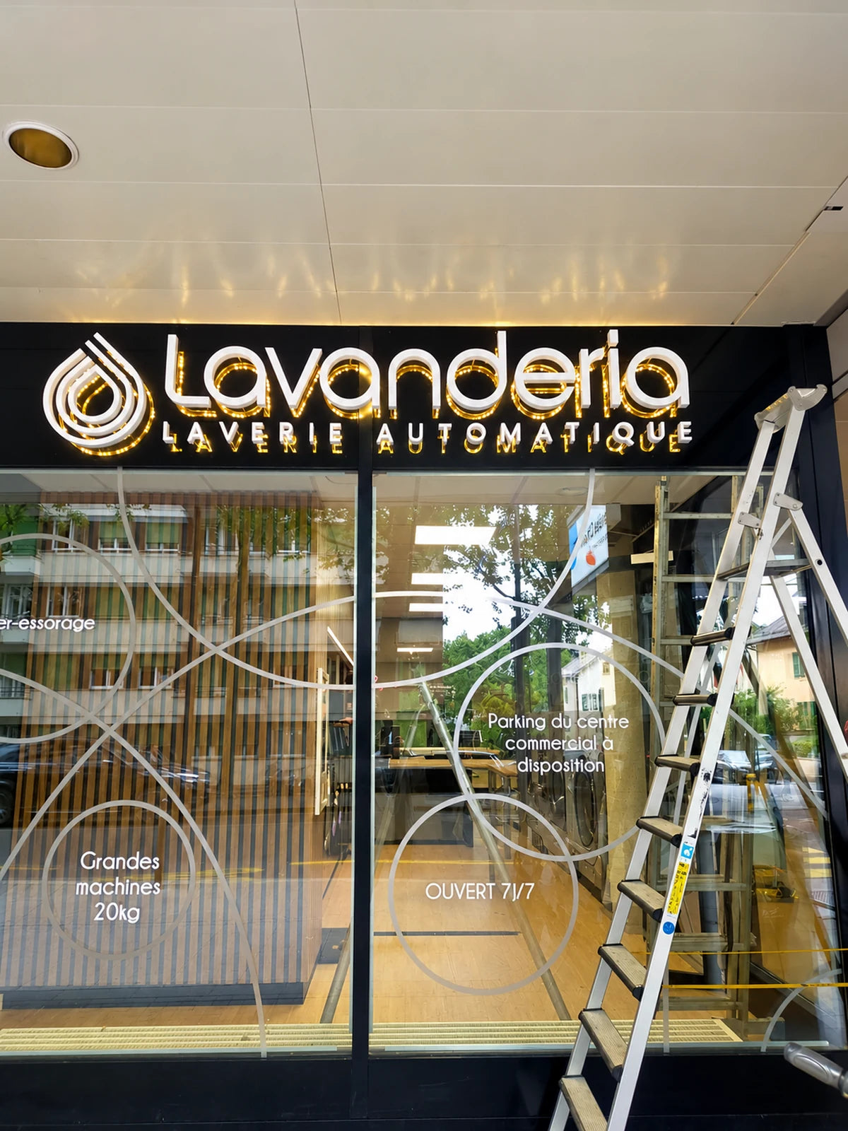 Montierte Lavanderia-Leuchtreklame an einer verglasten Fassade in Genf, mit weissen 3D-Buchstaben und einem Lichtkranz hinter dem Logo.