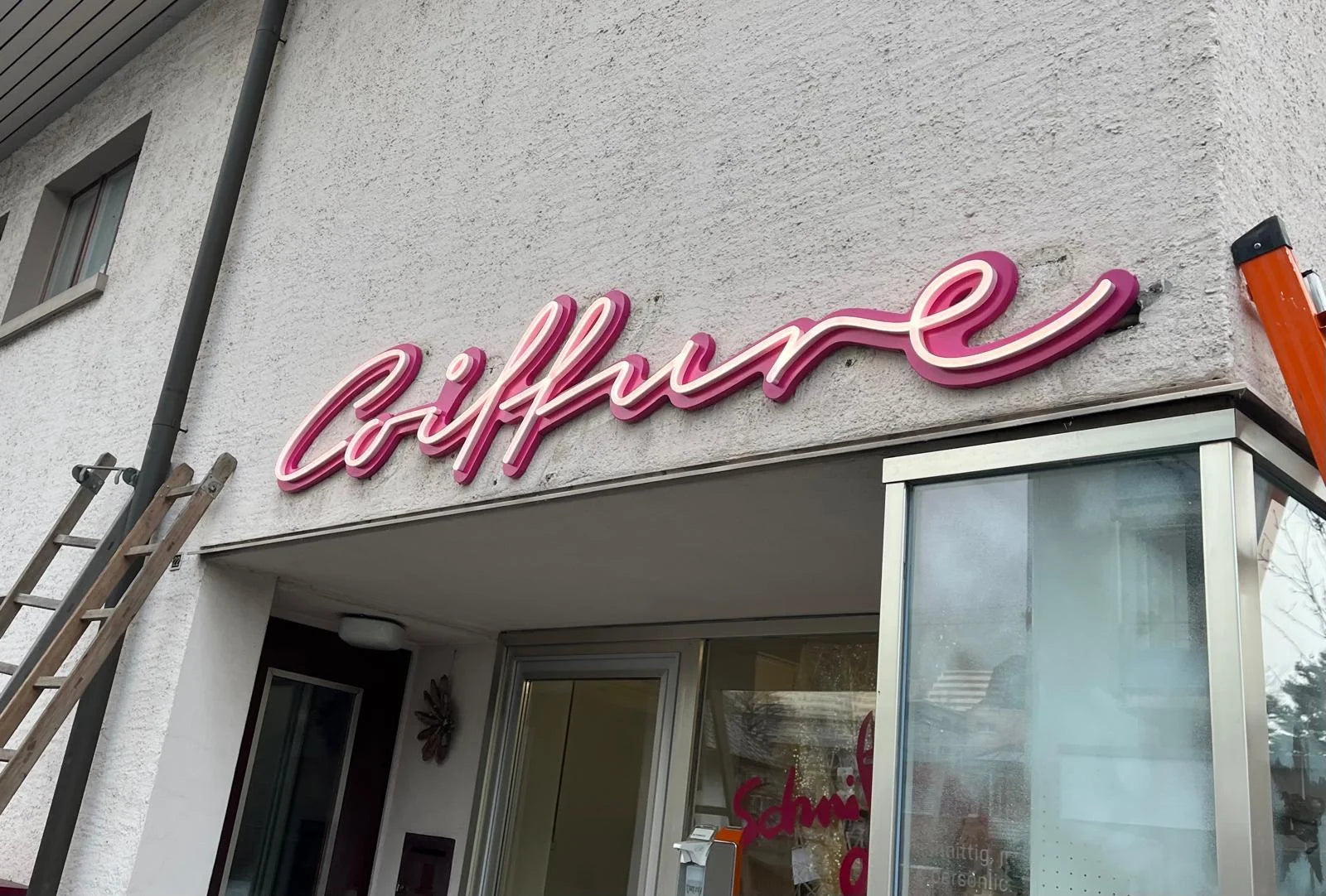 Coiffure-Leuchtschrift an der hell verputzten Salonfassade in Konolfingen, mit einer Leiter links und einer orangefarbenen Leiter rechts.