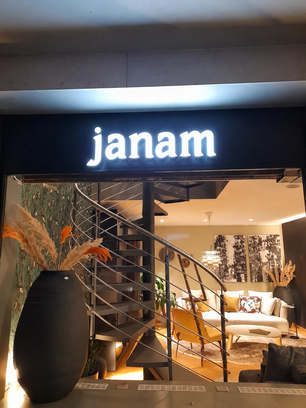 Illuminated janam sign installed on the shop’s black fascia in Lausanne, with the interior and staircase visible through the window.