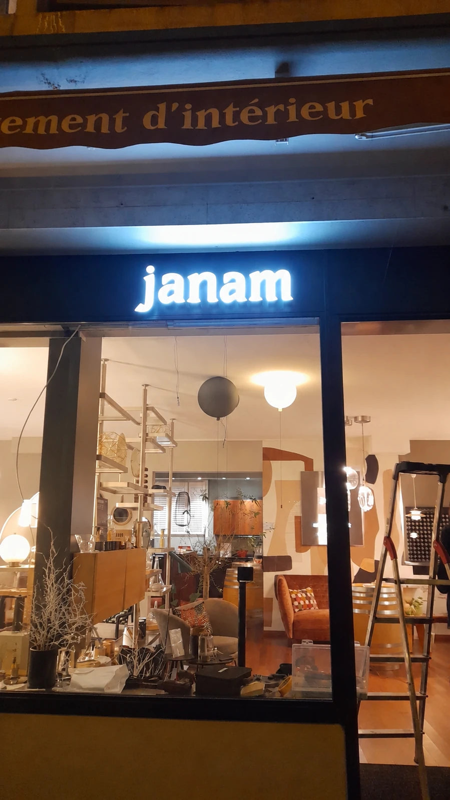 Illuminated janam sign on the shop fascia in Lausanne during the work on site, with a ladder visible inside.