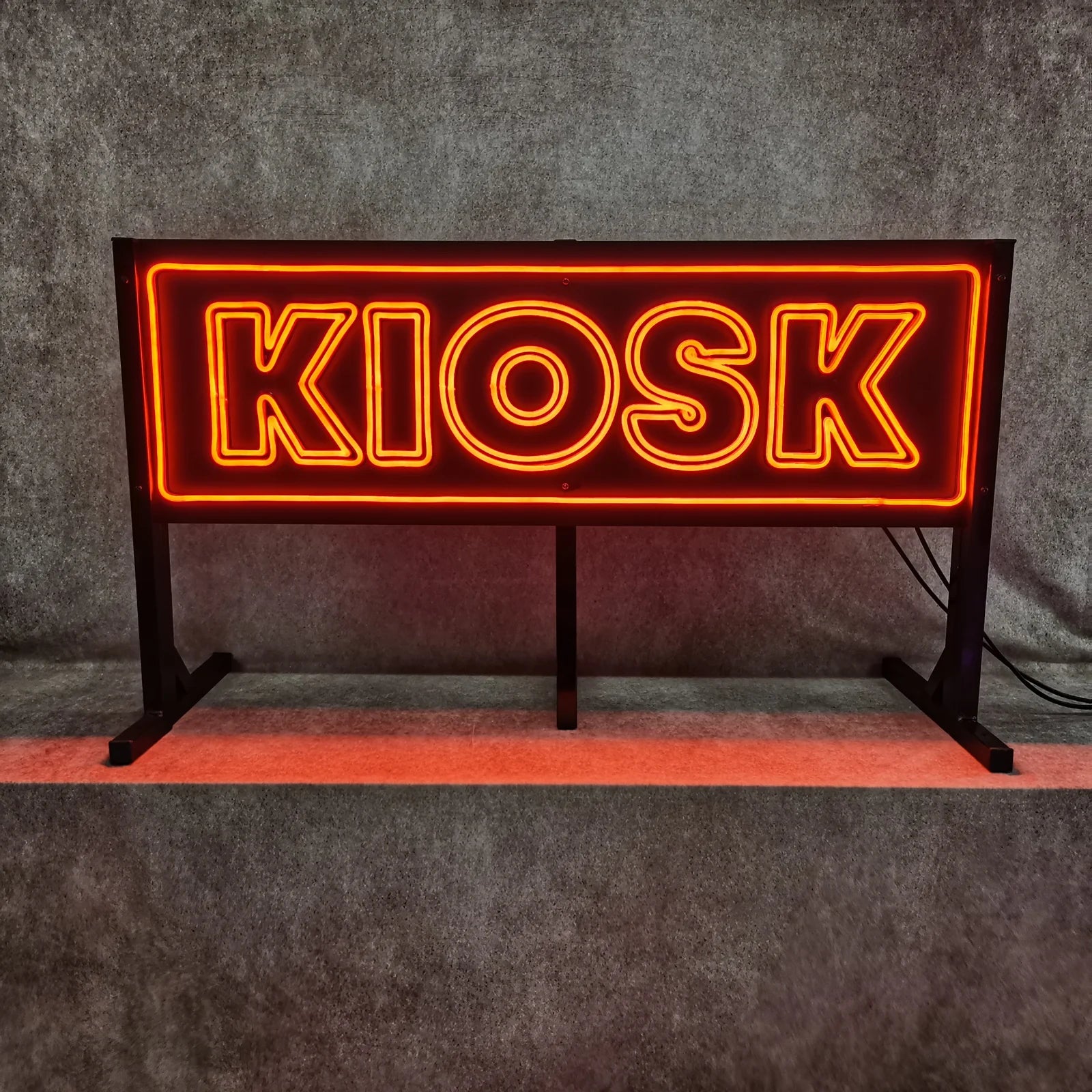 Leuchtendes KIOSK-Schild in der Werkstatt, frontal gesehen, mit orangefarbenen LED-Neonlinien und Leuchtrahmen auf dunklem Hintergrund.