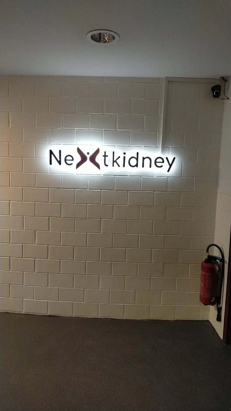 Indoor Nextkidney sign installed on a white wall in Lausanne, with a coloured logo, raised black lettering and a halo of backlighting.